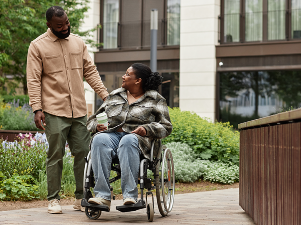A black couple gazes at one another, the woman is sat in a wheelchair with a coffee in her hand