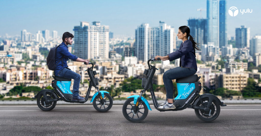 A man and a woman wearing smart clothing on electric scooters driving past each other. There is a city in the background