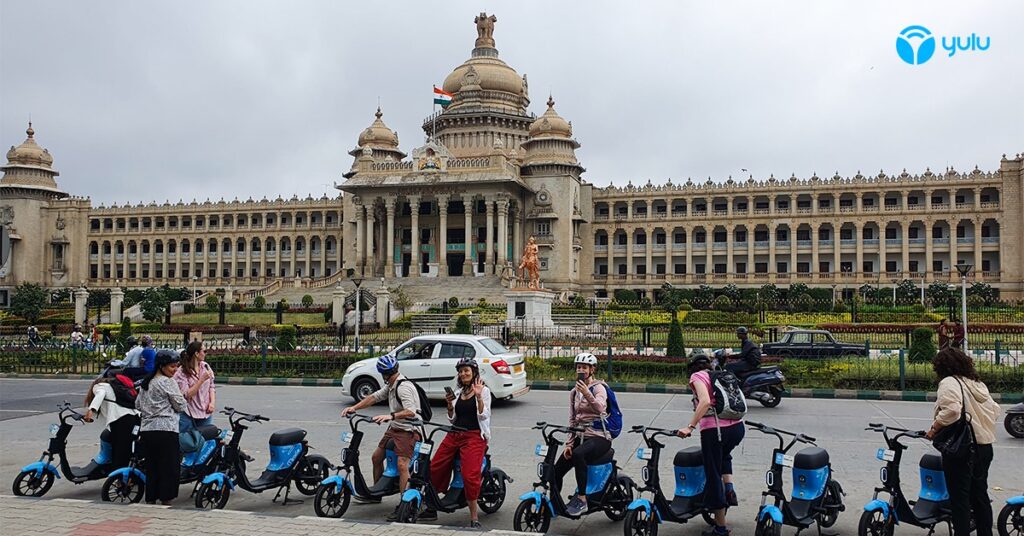Tourists on blue Yulu electric scooters pose in front of an ornate building