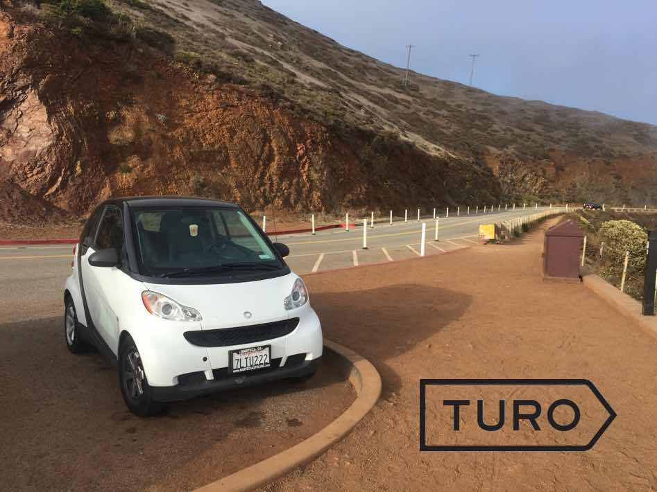 A small white electric vehicle parked at the side of the road. In the corner of the image is the TURO logo