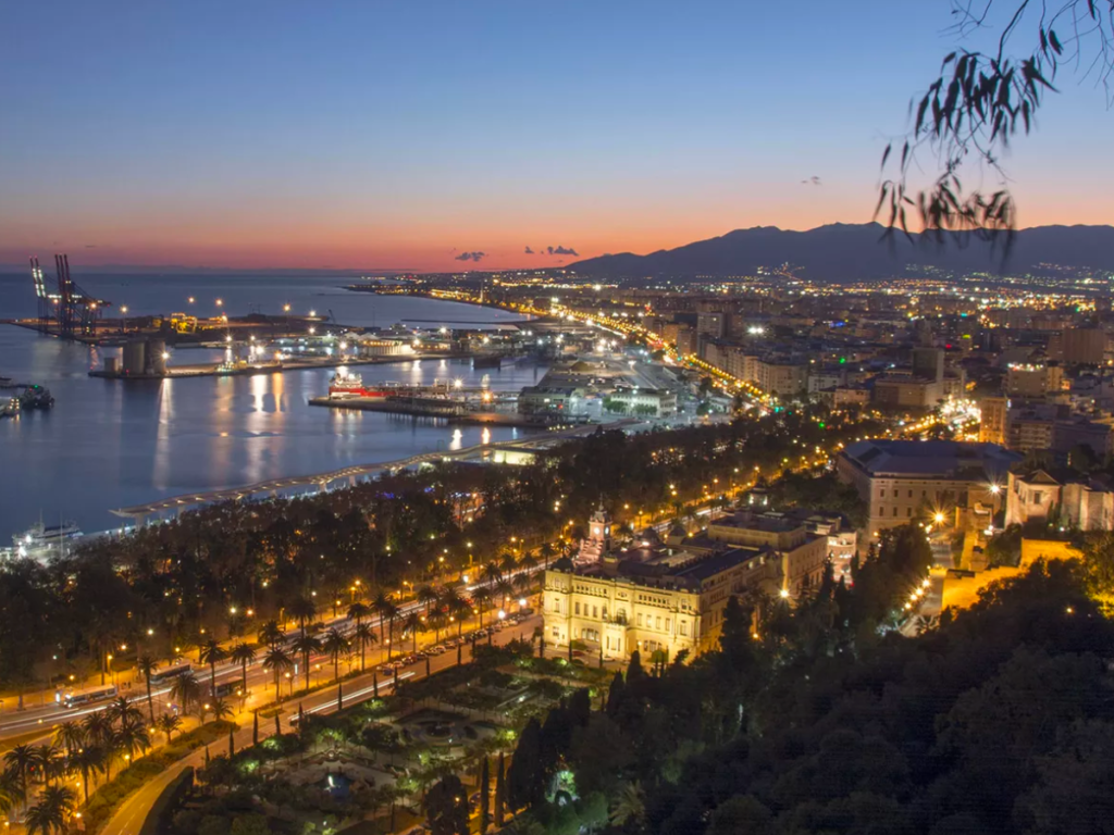 The city of Málaga at sunset. The city lights shine bright and the sky is a gradient from dark blue to orange
