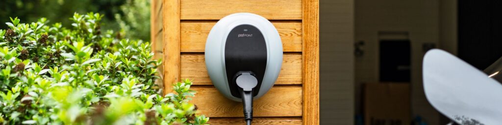 A pod point charger on a wooden garage beam next to a bush