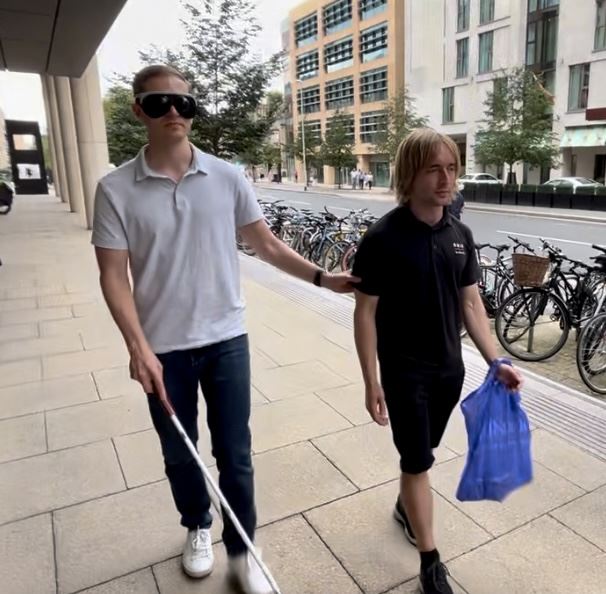 A person wearing a blindfold and using a walking stick is holding onto an RNIB employee as they're directed down a street