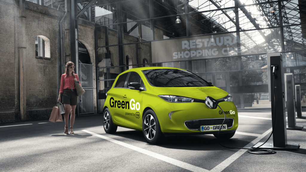 There is a green electric car with the words "GreenGo" written on the side. Its parked outside a modern shopping centre and there is a woman in pink walking towards it