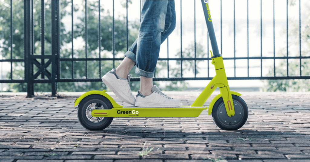 We see a persons legs from the knee down - they are wearing blue cropped jeans and white sneakers. The person is standing on a green electric scooter with the "GreenGo" logo written on the side