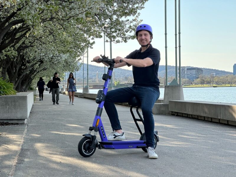 A man in a black shirt and jeans, wearing a purple bike helmet, is sat smiling on a purple beam scooter