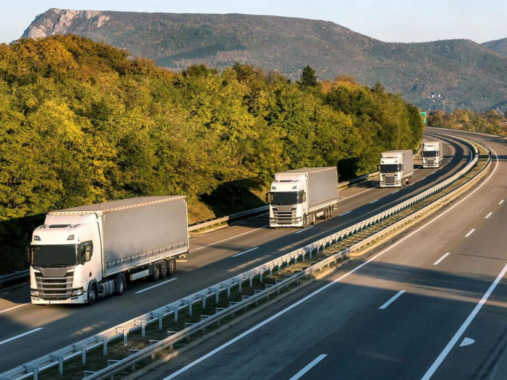 Four trucks driving on a highway. There are mountains behind them and large trees to the left side
