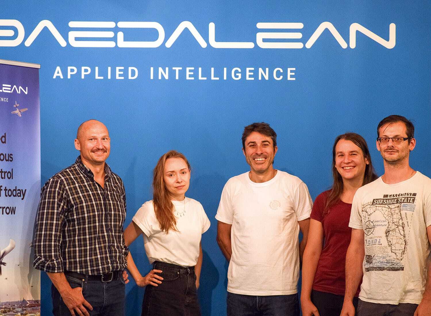 5 people, 3 men and 2 women, stood in front of a blue wall with the Daedalean logo