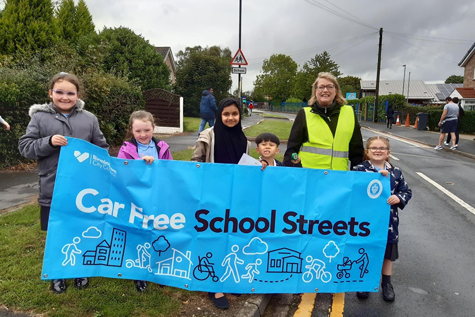 Children and staff members standing on a local road, holding a blue sign with the words 'Car-Free School Streets'