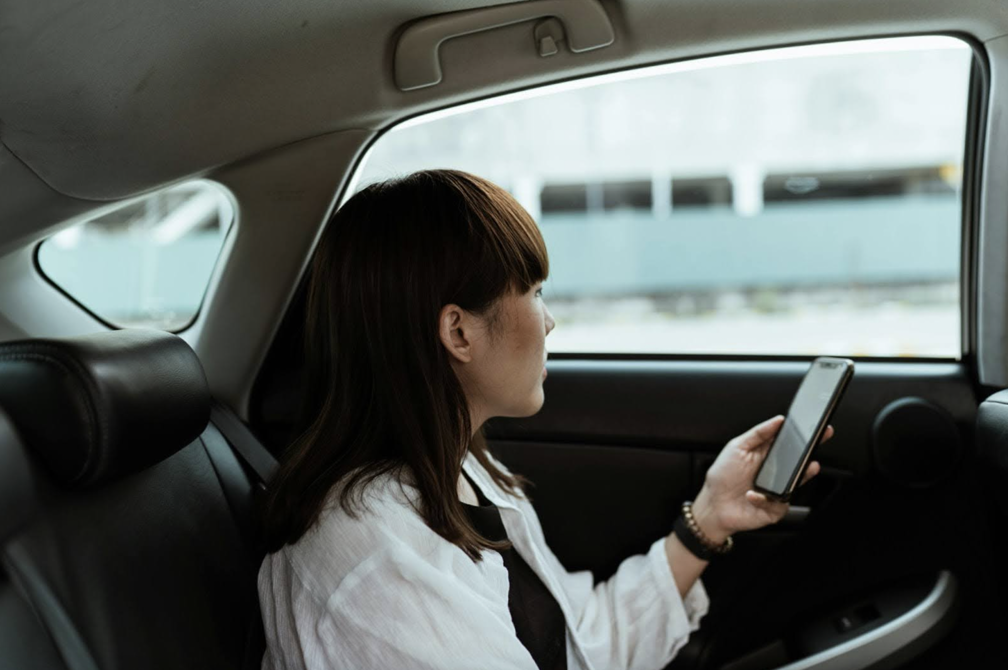 A woman sat in the back of a car, she is looking away from the camera out of the window, holding her smartphone in the left hand