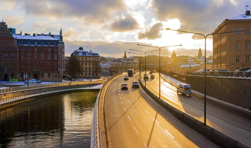 Scenic winter sunset over a busy highway in Stockholm, Sweden, with historic buildings, streetlights, and reflections on the water.