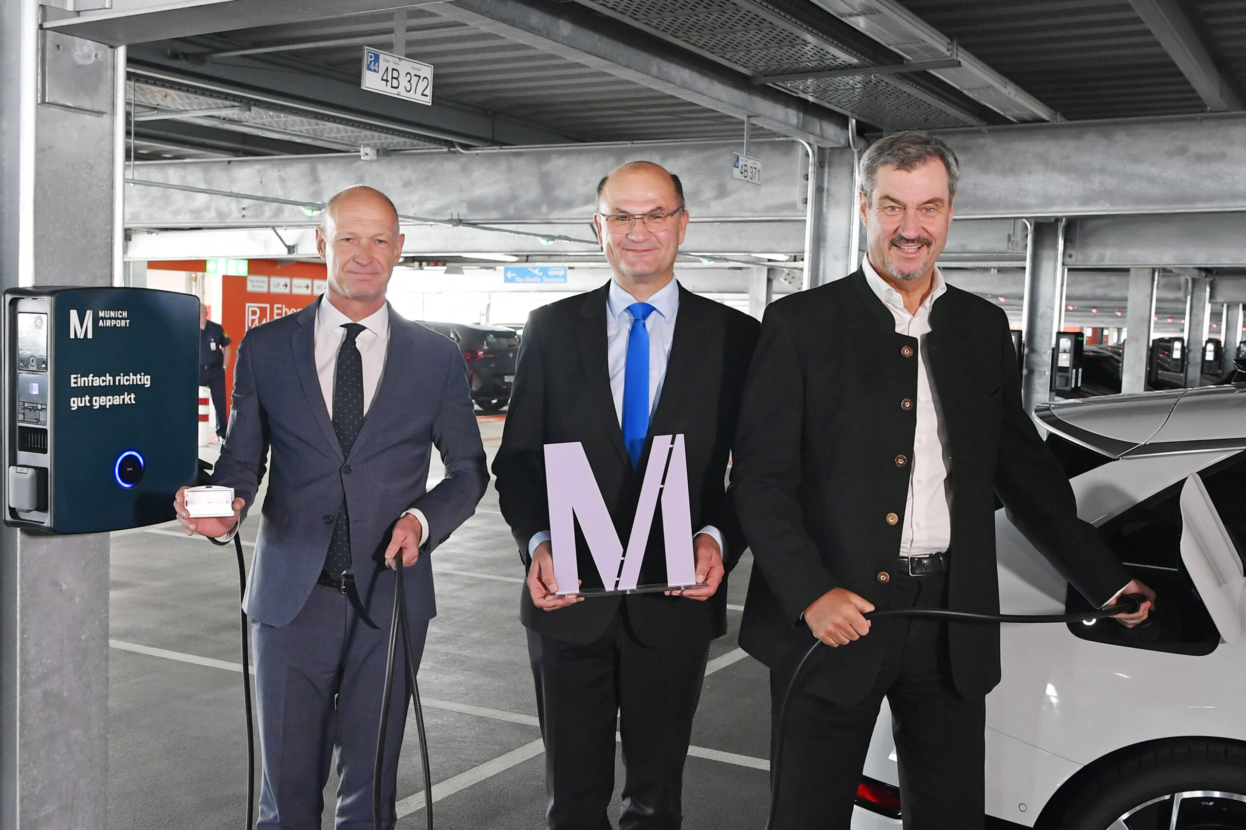Three men stood beside an electric car charging point, one is holding an M