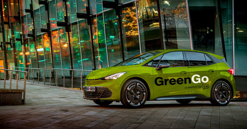 Lime-green CUPRA Born electric hatchback with GreenGo branding, parked on a paved plaza beside a glass building reflecting festive lights at dusk.