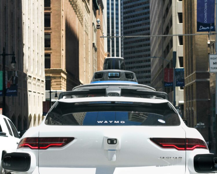 Rear view of a white Waymo autonomous Jaguar robotaxi with roof-mounted sensors driving along a city street