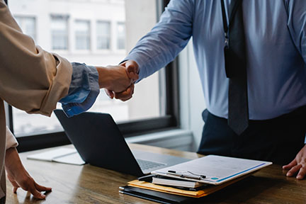 Two business partners shaking hands over a desk with a laptop and documents